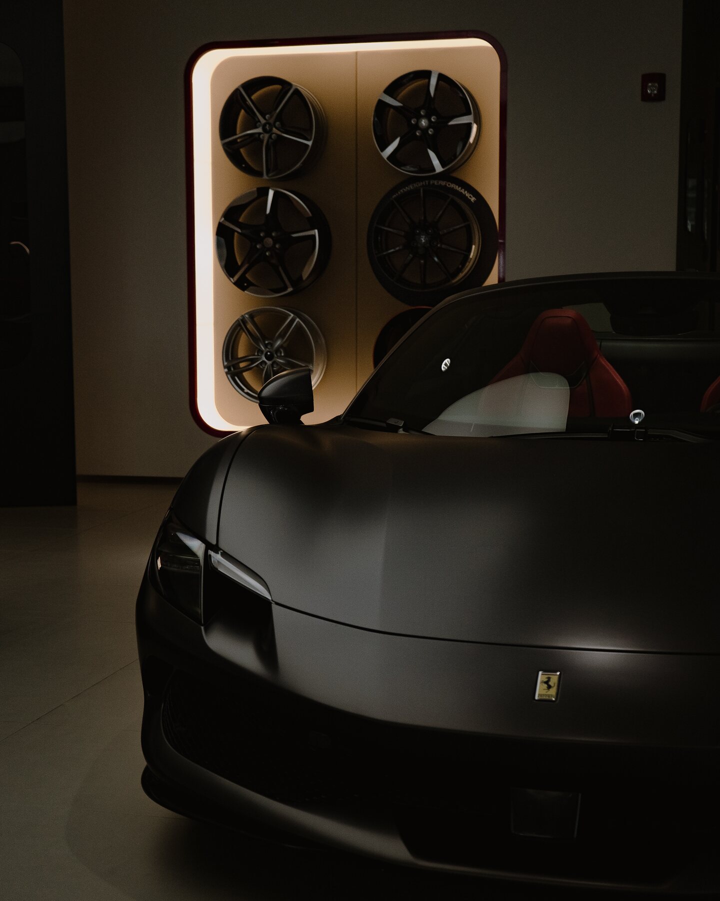 Matte gray Ferrari photographed beneath a wall display of wheels.