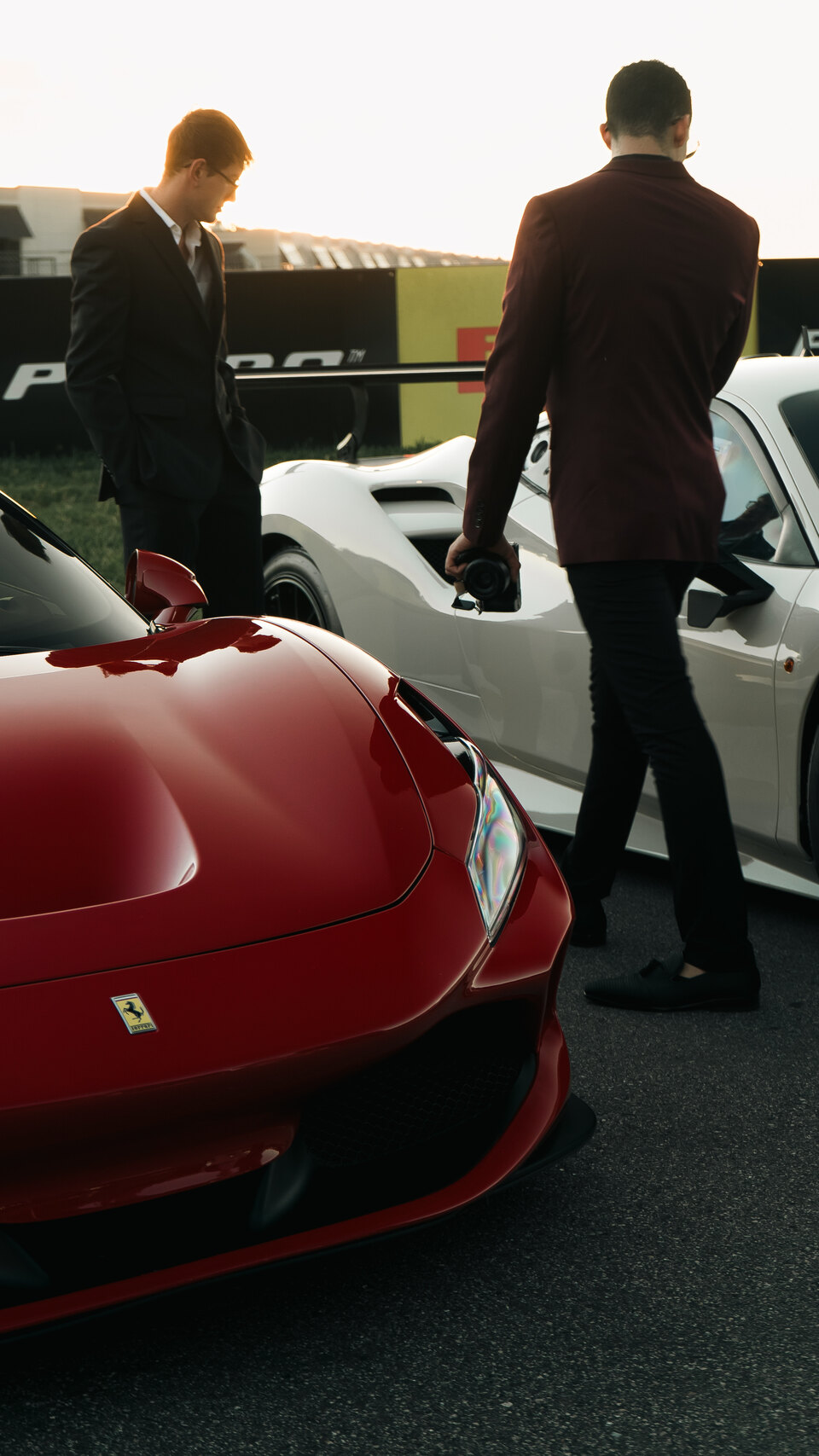 Well-dressed guests gather around exotic cars during a sunset automotive event.